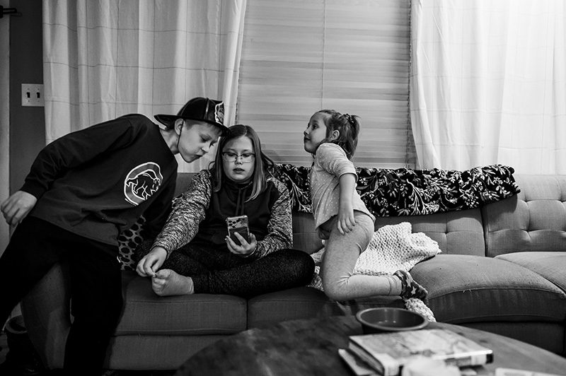 Black and white image of three young children on couch in living room Facetiming on phone by Denver photographer Elizabeth Osberg