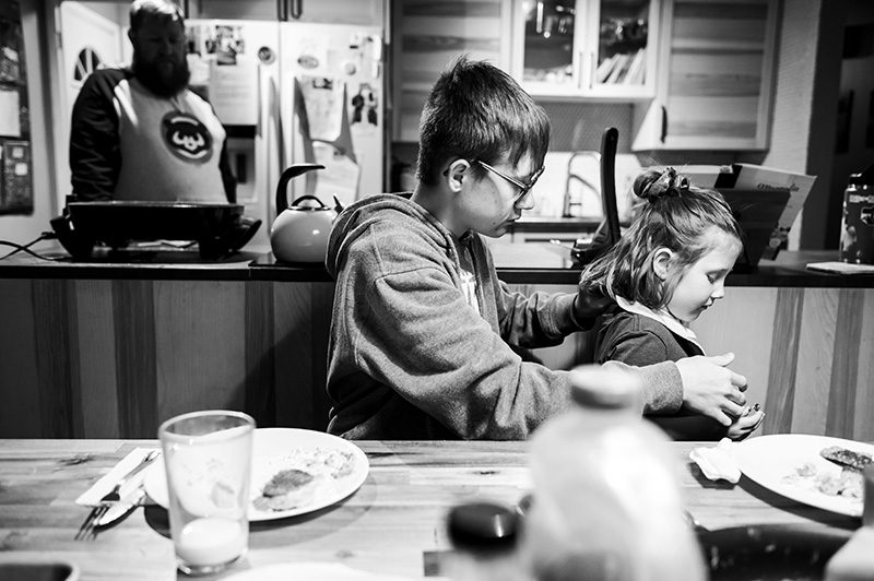 Black and white image of teenage brother helping preschool sister at the dinner table by Denver photographer Elizabeth Osberg