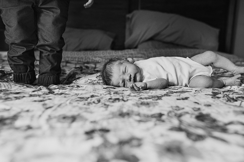 black and white image of baby sister lying on mom and dad's bed, brother standing very near on bed by denver, colorado photographer elizabeth osberg
