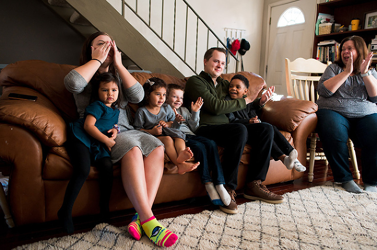 Family sitting on couch in living room of home. Mom crying tears of joy, dad and kids excitedly clapping at the close of an adoption by Denver, Colorado photographer Elizabeth Osberg