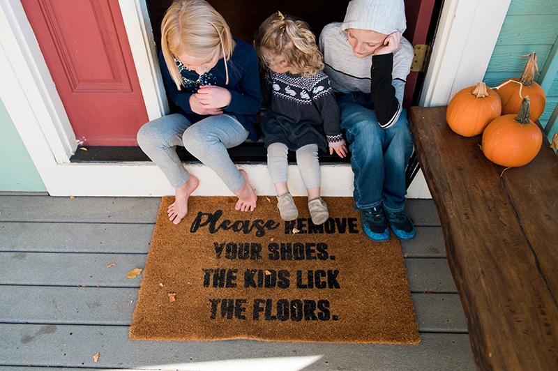Overhead view of three kids sitting on their doorstep with their feet on the door mat that reads, 